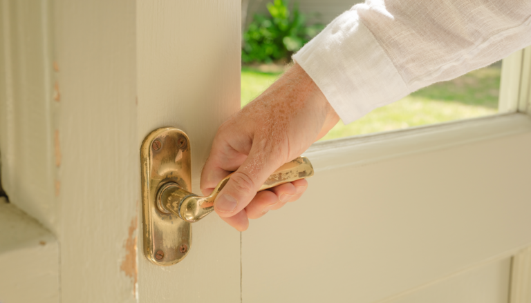 A hand holding a worn brass door handle beside a sunlit garden.