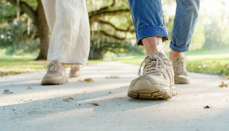 Two adults in trainers walking together along a sunlit, tree-lined sidewalk.